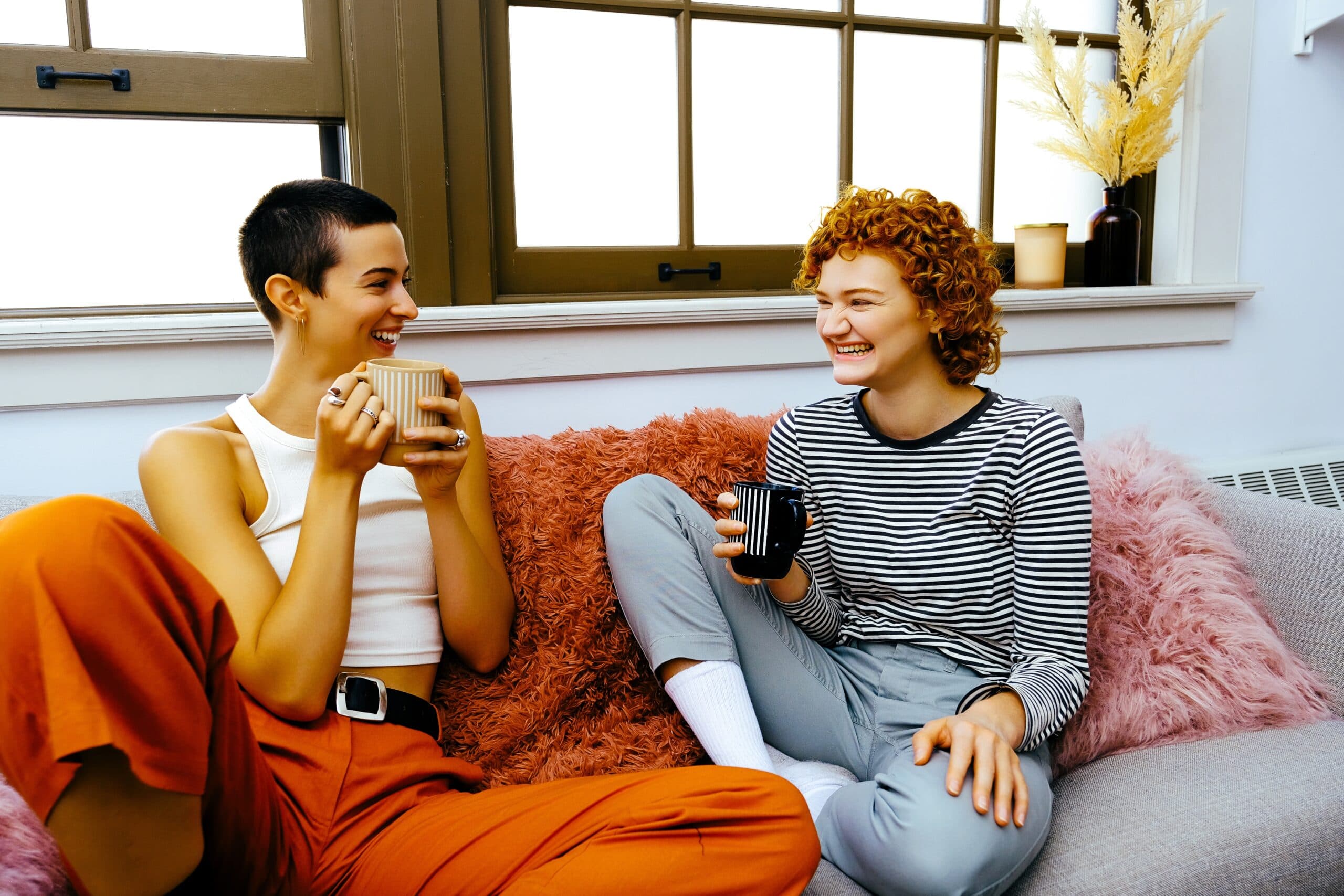 Two,Young,Women,Sitting,On,Couch,Holding,Mugs,,Smiling,And